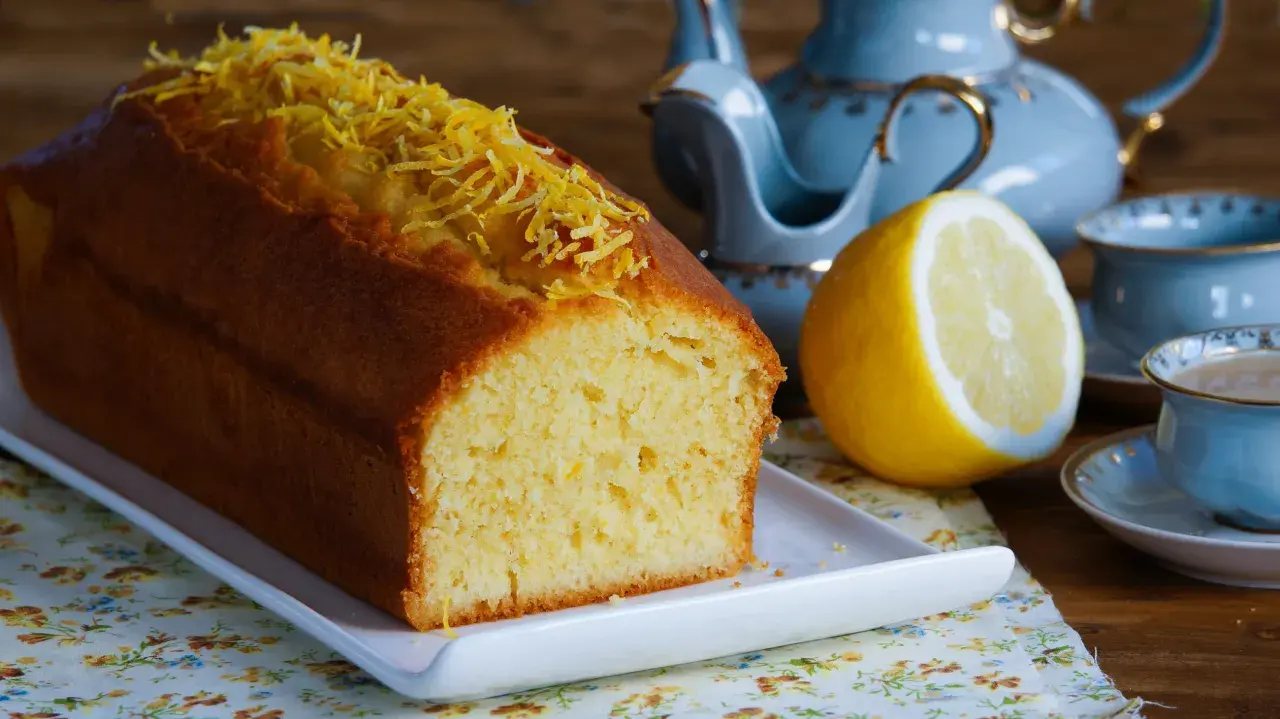 Puszysta babka piaskowa cytrynowa, ozdobiona skórką cytrynową, obok połówki cytryny i filiżanki herbaty.