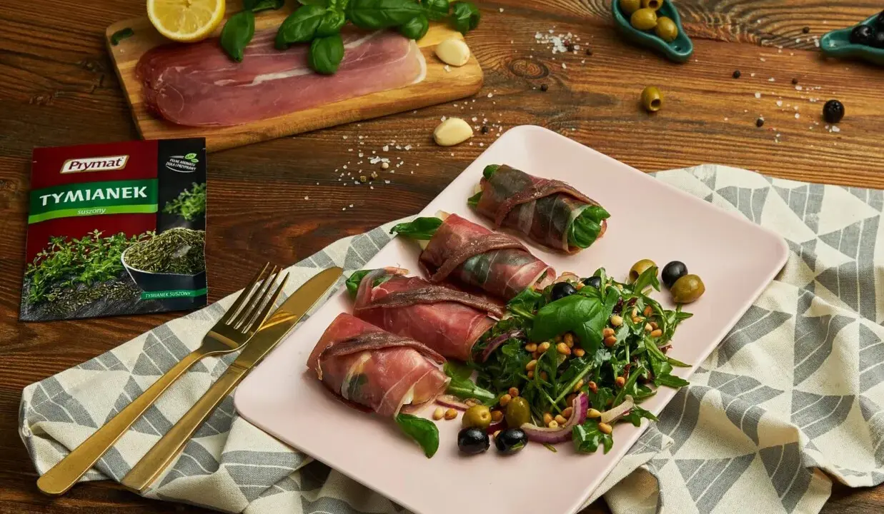 Roladki z szynką parmeńską i anchois, podane z rukolą, oliwkami i orzeszkami pinii. Obok talerza złote sztućce i opakowanie tymianku.