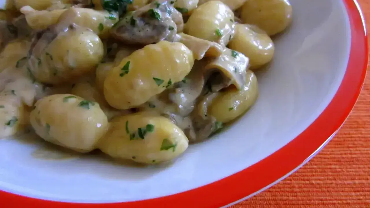 Pyszne gnocchi z sosem grzybowym i natką pietruszki, podane w białej misce z pomarańczowym rantem.