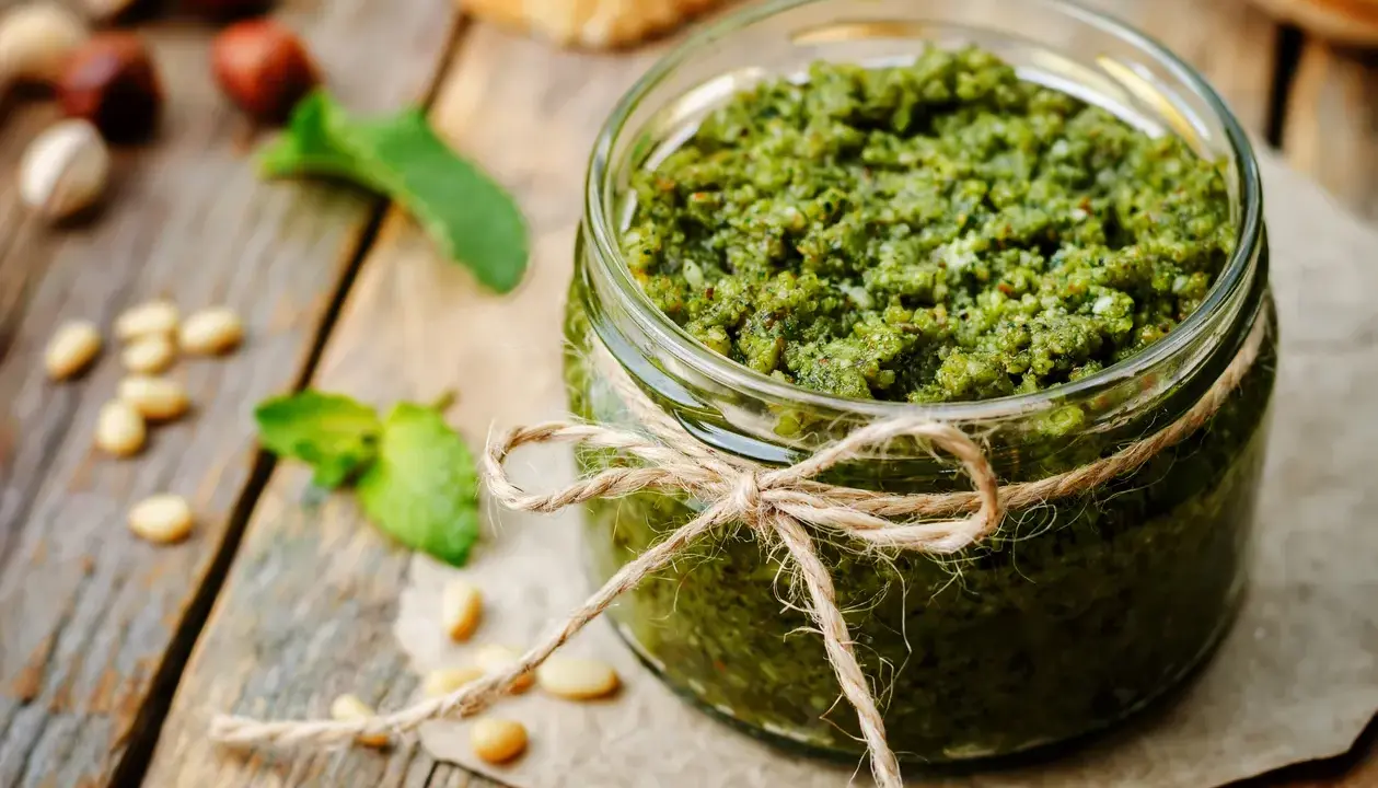 Słoik domowego pesto z bazylii, przewiązany sznurkiem. Obok orzeszki piniowe i listki bazylii.