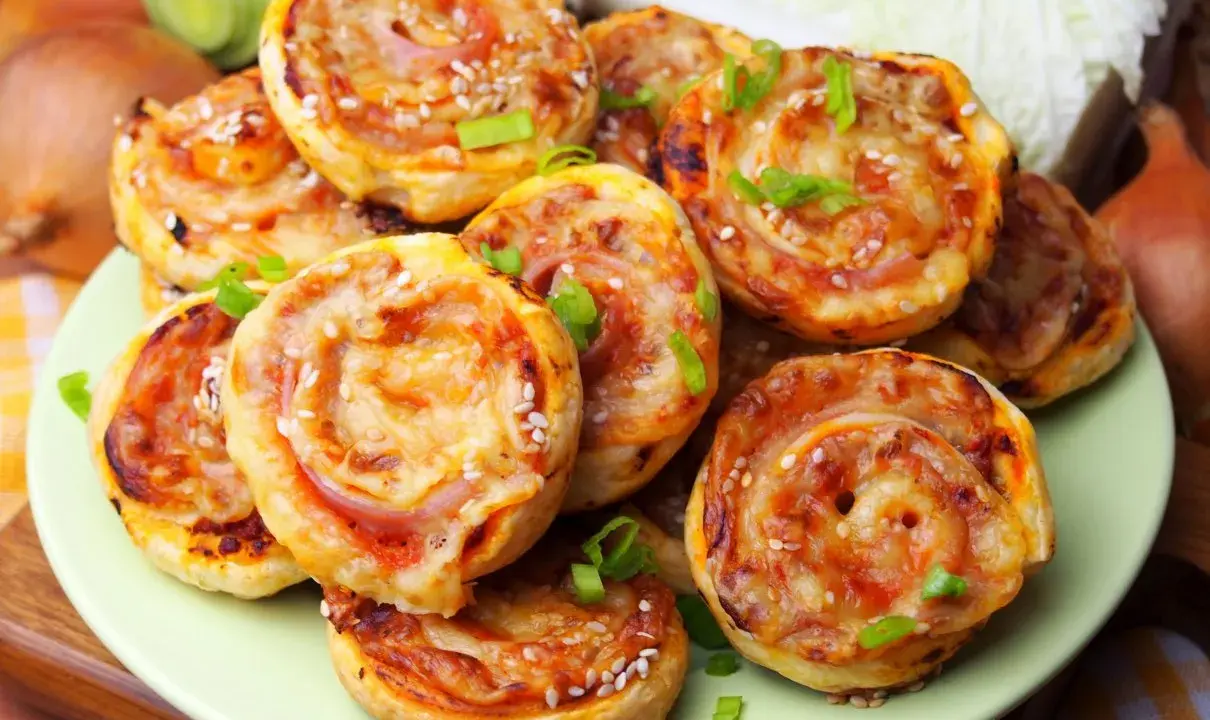 Pyszne pizzerinki z ciasta francuskiego, posypane sezamem i szczypiorkiem, ułożone na talerzu.