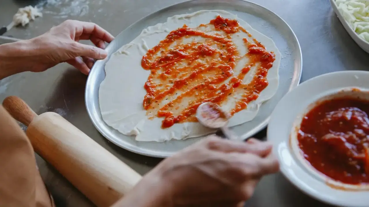 Ręce rozprowadzają pyszny sos do pizzy na cieście. Obok leży wałek i miseczka z sosem.