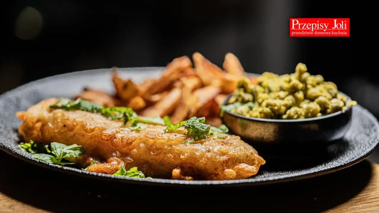 Chrupiący fish and chips z zielonym groszkiem i świeżą miętą na czarnym talerzu.
