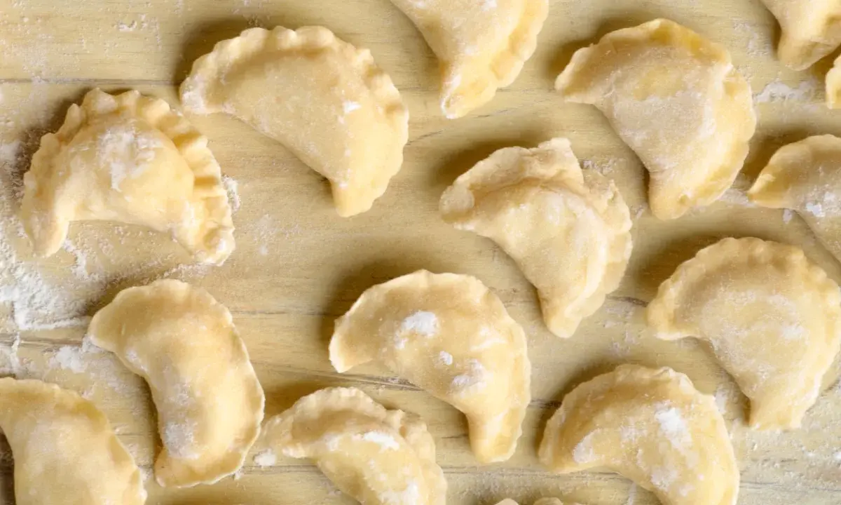 Surowe pierogi z farszem na pierogi ruskie, ułożone na drewnianym blacie, posypane mąką.