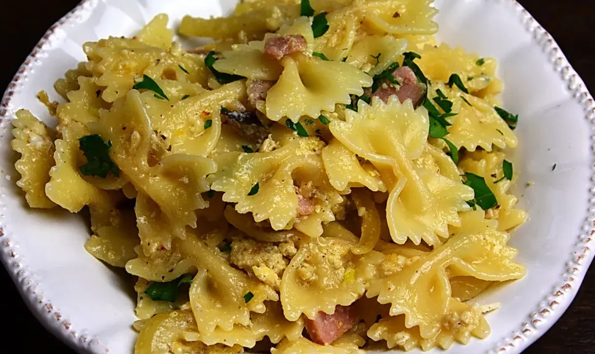 Pyszny makaron farfalle z jajkiem, kawałkami szynki i posiekaną natką pietruszki, podany w białej misce.