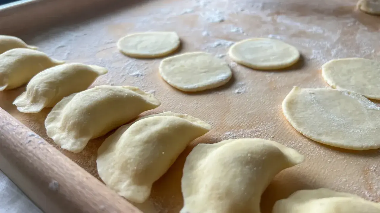 Gotowe do lepienia pierogi ruskie. Na drewnianej desce leżą okrągłe placki ciasta i uformowane pierożki.