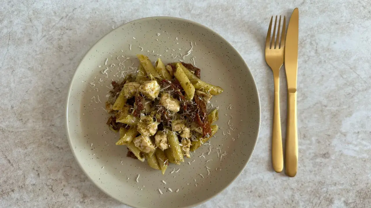Penne z kurczakiem i suszonymi pomidorami, polane aromatycznym makaronem z pesto ze słoika, posypane parmezanem. Obok złote sztućce.