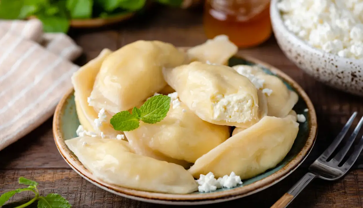 Talerz pełen pysznych, domowych pierogów z serem, udekorowany miętą. Obok miseczka z twarogiem i słoiczek miodu.