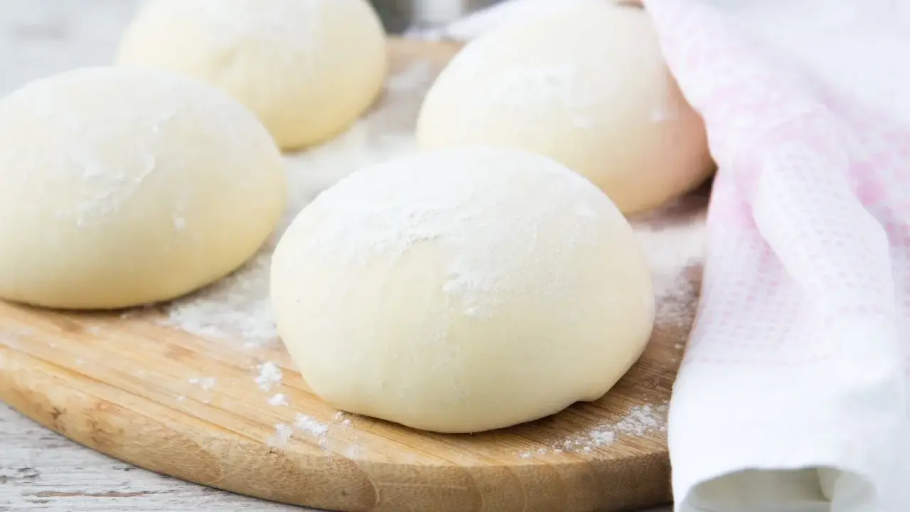 Kulki ciasta na spód do pizzy, posypane mąką, leżą na drewnianej desce. Obok leży różowy ręcznik.