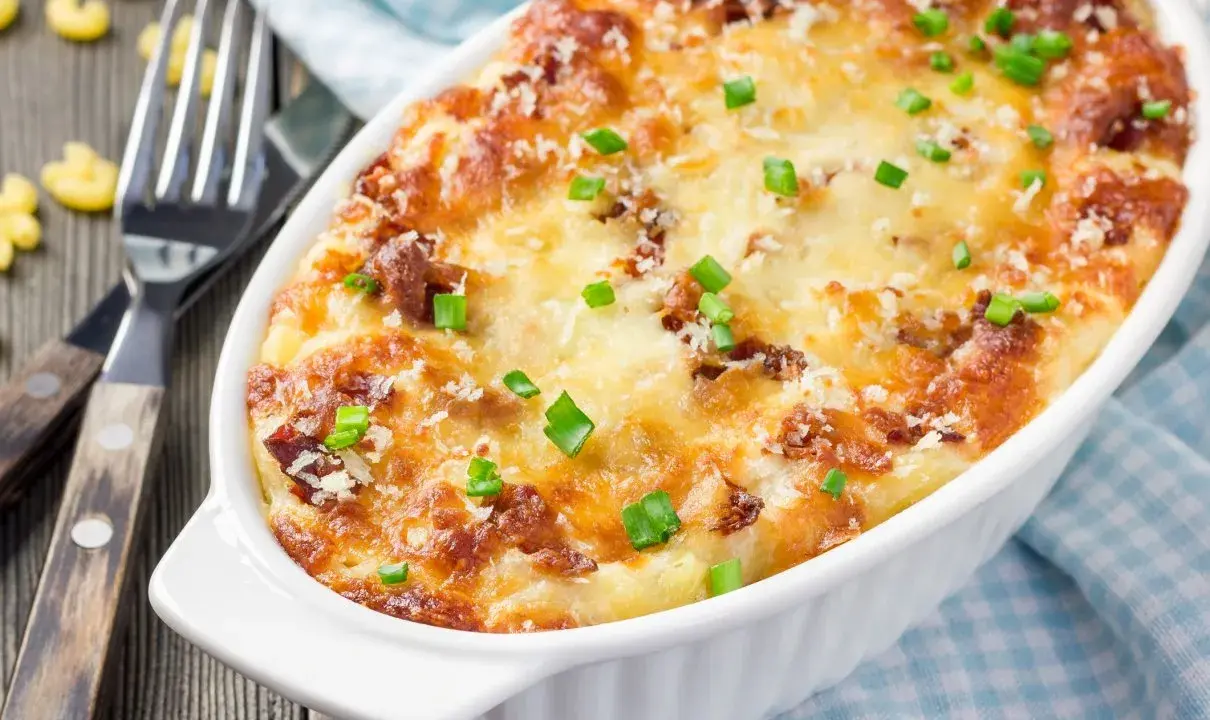 Pyszne, zapiekane mac and cheese w białym naczyniu, posypane szczypiorkiem i parmezanem. Obok widelce i luźne makarony.