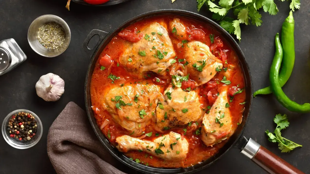 Pyszny kurczak w sosie pomidorowym, duszony z cebulą i przyprawami, udekorowany natką pietruszki. Obok zielona papryczka chili i pomidory.