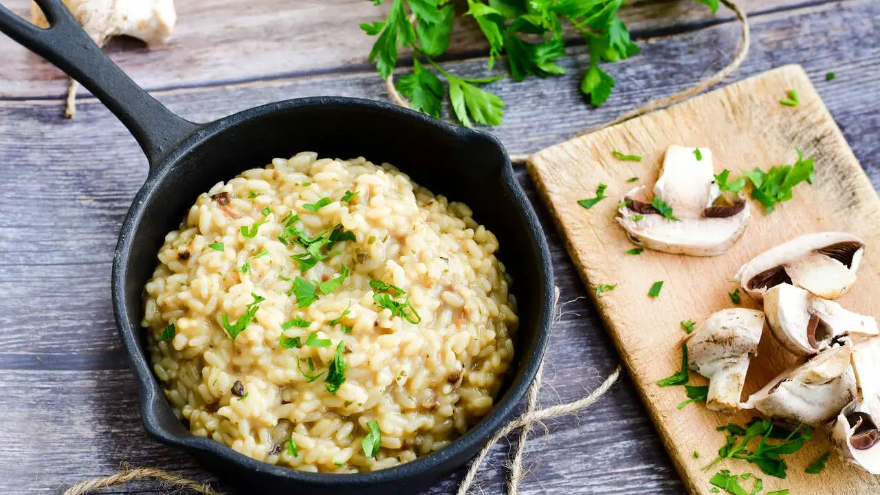 Kremowe risotto z warzywami, posypane natką pietruszki, obok świeże pieczarki na desce.