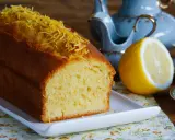Puszysta babka piaskowa cytrynowa, ozdobiona skórką cytrynową, obok połówki cytryny i filiżanki herbaty.