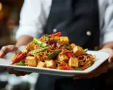 Talerz pełen aromatycznego makaronu z tofu, chrupiącymi warzywami i sosem. Danie prezentuje kucharz.