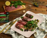 Roladki z szynką parmeńską i anchois, podane z rukolą, oliwkami i orzeszkami pinii. Obok talerza złote sztućce i opakowanie tymianku.