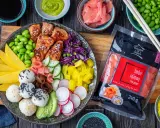 Kolorowy bowl z lososiem, ryżowymi kulkami, mango, awokado, edamame i marynowanym imbirem. Obok opakowanie różowego imbiru.