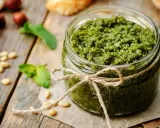 Słoik domowego pesto z bazylii, przewiązany sznurkiem. Obok orzeszki piniowe i listki bazylii.