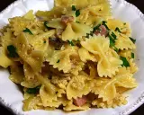 Pyszny makaron farfalle z jajkiem, kawałkami szynki i posiekaną natką pietruszki, podany w białej misce.