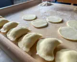 Gotowe do lepienia pierogi ruskie. Na drewnianej desce leżą okrągłe placki ciasta i uformowane pierożki.