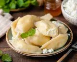 Talerz pełen pysznych, domowych pierogów z serem, udekorowany miętą. Obok miseczka z twarogiem i słoiczek miodu.