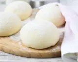 Kulki ciasta na spód do pizzy, posypane mąką, leżą na drewnianej desce. Obok leży różowy ręcznik.
