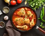 Pyszny kurczak w sosie pomidorowym, duszony z cebulą i przyprawami, udekorowany natką pietruszki. Obok zielona papryczka chili i pomidory.