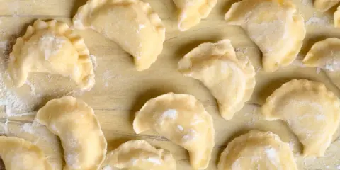 Surowe pierogi z farszem na pierogi ruskie, ułożone na drewnianym blacie, posypane mąką.
