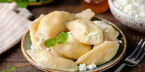 Pyszne pierogi z serem na słodko, posypane twarogiem i udekorowane miętą. Obok słoiczek miodu i miseczka z twarogiem.