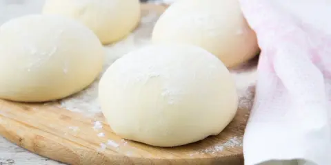 Kulki ciasta na spód do pizzy, posypane mąką, leżą na drewnianej desce. Obok leży różowy ręcznik.