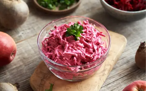 Pyszne buraczki z chrzanem w szklanej misce, ozdobione natką pietruszki. Obok surowe buraki i jabłko.