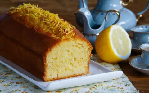 Puszysta babka piaskowa cytrynowa, ozdobiona skórką cytrynową, obok połówki cytryny i filiżanki herbaty.