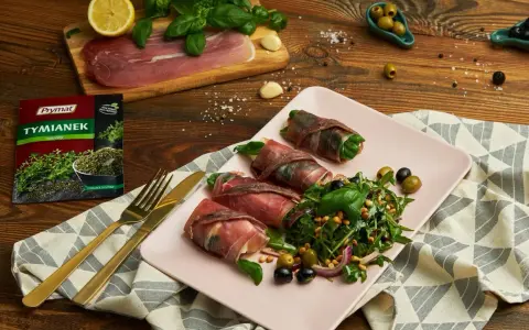 Roladki z szynką parmeńską i anchois, podane z rukolą, oliwkami i orzeszkami pinii. Obok talerza złote sztućce i opakowanie tymianku.