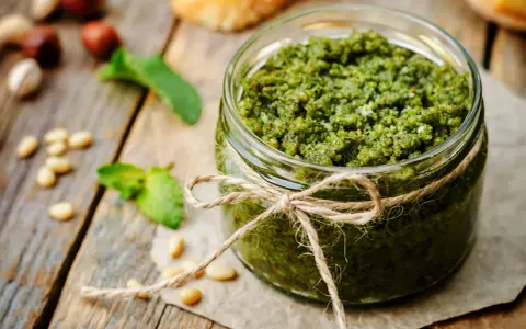 Słoik domowego pesto z bazylii, przewiązany sznurkiem. Obok orzeszki piniowe i listki bazylii.