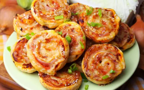 Pyszne pizzerinki z ciasta francuskiego, posypane sezamem i szczypiorkiem, ułożone na talerzu.