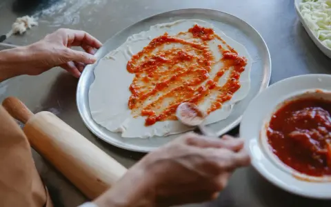 Ręce rozprowadzają pyszny sos do pizzy na cieście. Obok leży wałek i miseczka z sosem.