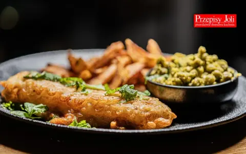 Chrupiący fish and chips z zielonym groszkiem i świeżą miętą na czarnym talerzu.