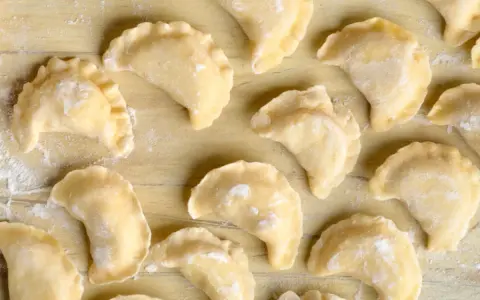 Surowe pierogi z farszem na pierogi ruskie, ułożone na drewnianym blacie, posypane mąką.