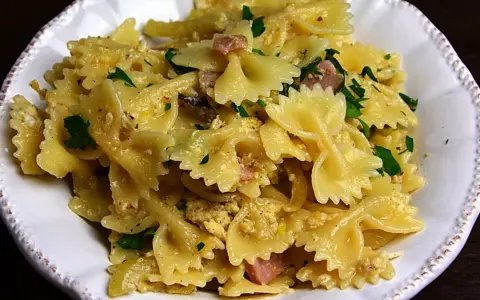Pyszny makaron farfalle z jajkiem, kawałkami szynki i posiekaną natką pietruszki, podany w białej misce.