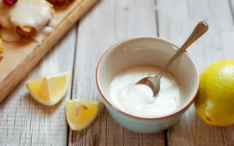 Gęsty, biały lukier do cynamonek w miseczce, obok cytryny i kawałków cytryny.