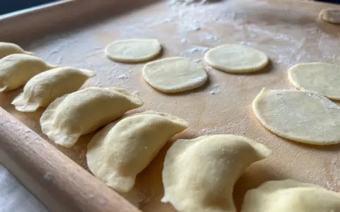 Gotowe do lepienia pierogi ruskie. Na drewnianej desce leżą okrągłe placki ciasta i uformowane pierożki.