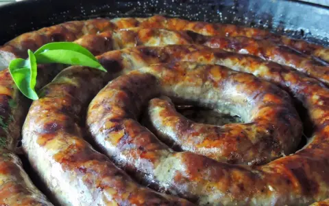 Złocista, pieczona biała kiełbasa w kształcie spirali, skropiona tłuszczem, z zielonym listkiem.