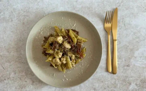 Penne z kurczakiem i suszonymi pomidorami, polane aromatycznym makaronem z pesto ze słoika, posypane parmezanem. Obok złote sztućce.