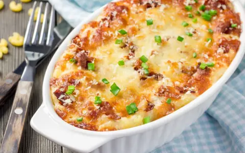 Pyszne, zapiekane mac and cheese w białym naczyniu, posypane szczypiorkiem i parmezanem. Obok widelce i luźne makarony.