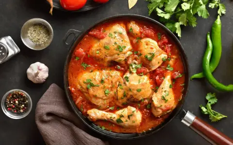 Pyszny kurczak w sosie pomidorowym, duszony z cebulą i przyprawami, udekorowany natką pietruszki. Obok zielona papryczka chili i pomidory.