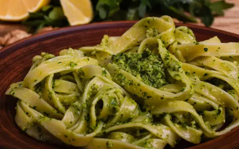 Tagliatelle z aromatycznym pesto z rukoli, podane na brązowym talerzu, z cytryną i orzechami w tle.