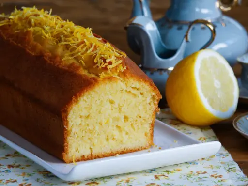 Puszysta babka piaskowa cytrynowa, ozdobiona skórką cytrynową, obok połówki cytryny i filiżanki herbaty.