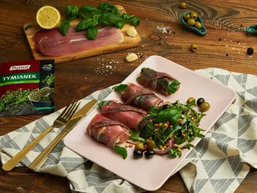 Roladki z szynką parmeńską i anchois, podane z rukolą, oliwkami i orzeszkami pinii. Obok talerza złote sztućce i opakowanie tymianku.