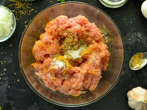Mielone mięso na kebab z przyprawami w szklanej misce. Obok posiekana cebula, czosnek i łyżeczka z przyprawami.