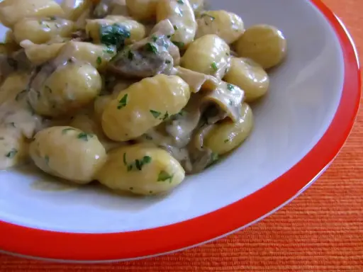 Pyszne gnocchi z sosem grzybowym i natką pietruszki, podane w białej misce z pomarańczowym rantem.