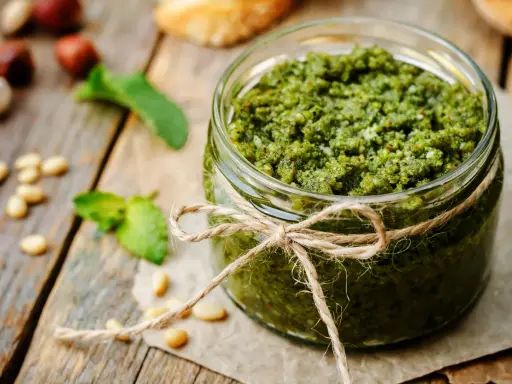 Słoik domowego pesto z bazylii, przewiązany sznurkiem. Obok orzeszki piniowe i listki bazylii.
